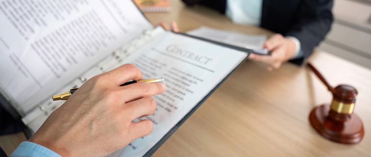 Two people in business attire sit at a desk reviewing a contract; one holds a pen and binder, while the other holds papers. A judges gavel is visible on the table.